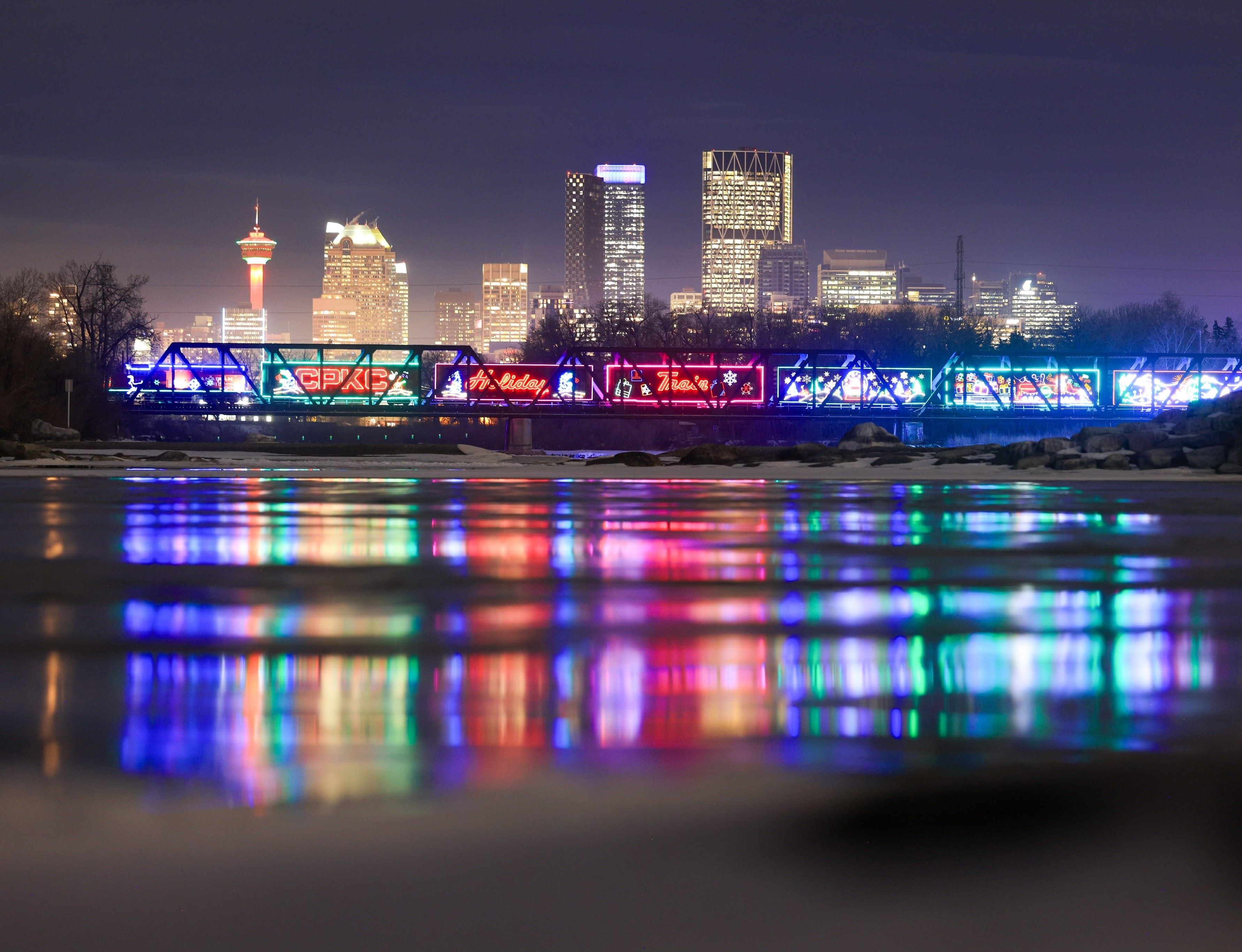 CPKC Holiday Train reflecting into the water as it travels on a bridge passing by a city in the background
